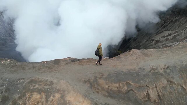 Aerial view of woman walking along the rim of Mount Bromo volcano crater surrounded by rising steam in East Java, Indonesia. Dramatic travel and adventure scene showcasing the raw power of nature.