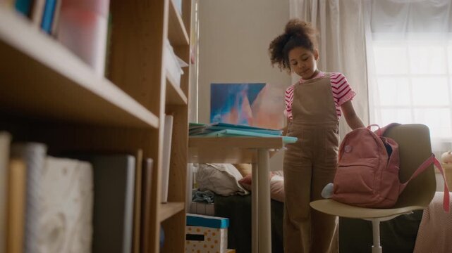 African American child packing school supplies into pink backpack in bedroom, preparing for school day. Ideal for back-to-school marketing campaigns, parenting promos, and educational content