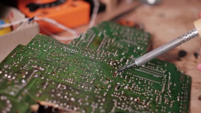 Technician hand precisely guides a soldering iron to melt solder onto connections of a green circuit board. The smoke rising from the board highlights manual assembly and repair.