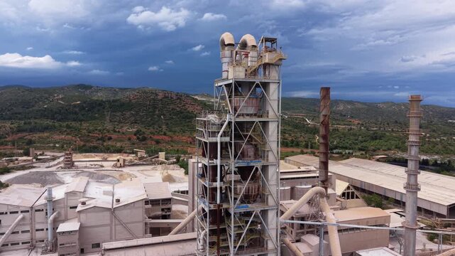 Large cement manufacturing facility featuring a multi-stage cyclone preheater tower, rotary kiln, steel framework structures, exhaust stacks, and industrial buildings