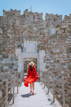 Young woman in a flowing red dress and hat walking towards the ancient entrance of Bodrum Castle on a sunny day
