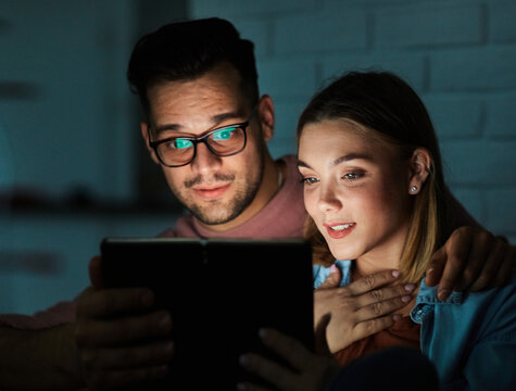 Portrait of young happy couple using tablet computer at night, using modern technology having fun with glowing screen in dark office or at home, watching tv together at home. Shot of a couple resting 