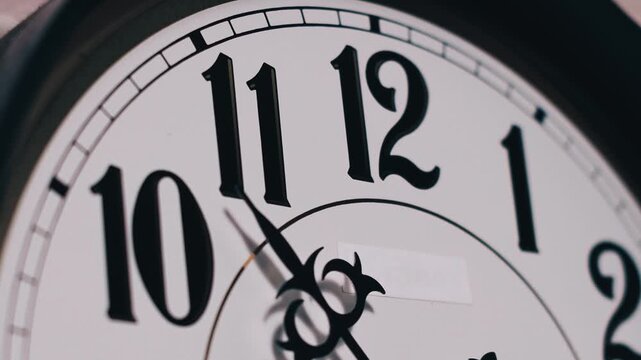 Macro time lapse shows analog clock hands racing over a clean white face with black numerals. The rapid motion marks time passage against the classic dial background.