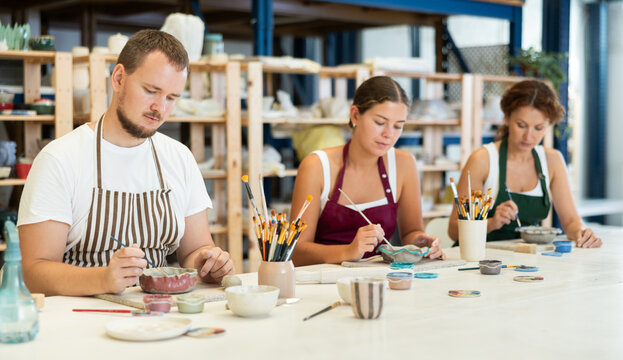 Three pottery masters work together on ceramic tableware, enjoying creative process as they paint mugs in the workshop