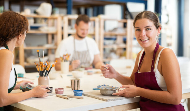 In pottery workshop, two girls and guy are sitting at table, busy coloring products made of raw clay. Amateurs are busy painting clay plates at master class