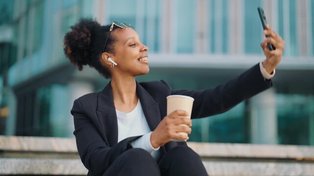 Cheerful black woman taking selfies by mobile phone when drinking coffee in park . Young adult african american lady in business suit photographing herself by smartphone, good mood and positivity