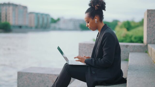 Freelancer woman working with laptop on river embankment in city, online job. African american lady surfing internet by portable computer with free wifi connection, female startup owner outdoors