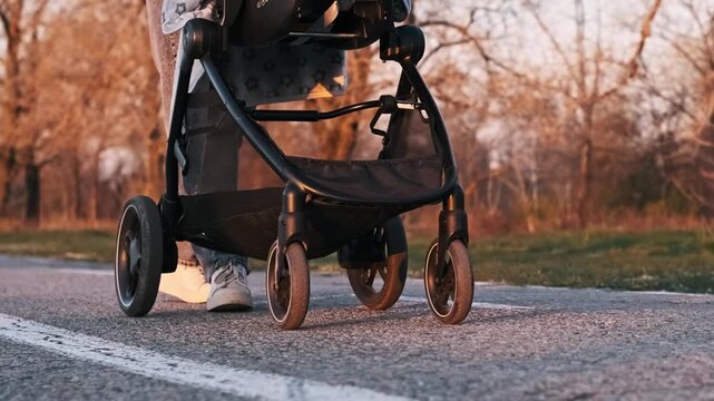 Ground level close-up of a parent in jeans and white sneakers pushing a baby stroller on a cracked asphalt park path. Warm sunset light illuminates the feet and wheel in motion.