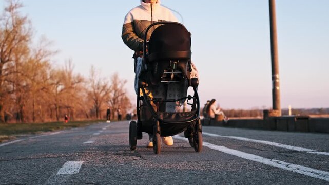 Low angle view shows a young mother in sunglasses walking towards the camera pushing a black baby stroller. The sunset light illuminates the asphalt path in a park setting.