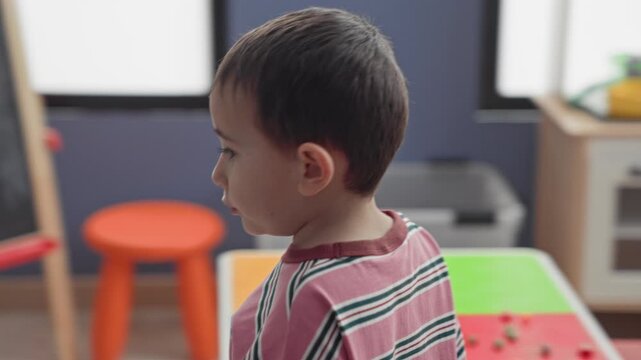 Boy touches his head, walks past bowling pins and reaches toward toy shelf full of plush and blocks in a building; simple childhood curiosity.