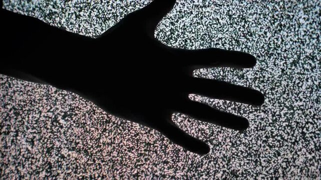 A close-up shot features a dark hand silhouette entering the frame from the left and moving horizontally across the flickering black and white static of an old television screen display.