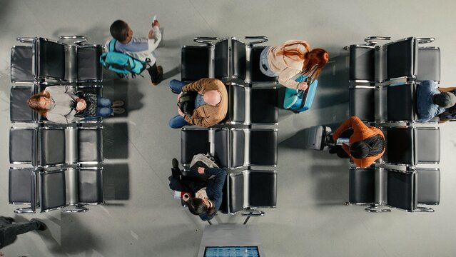 Travelers with trolleys waiting to do airport checkin, top down view, people walking in airplane terminal with baggage, passing by electronic flight timetable announcement board, high angle shot.