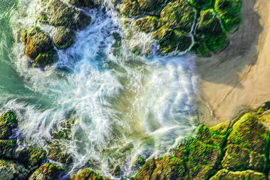 Aerial view of mossy rocks on the shore of Kingscliff Beach with white crashing waves and golden sand in Kingscliff, New South Wales, Australia.