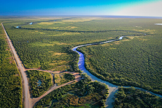 Aerial view of a winding river flowing through the vast green landscape of the outback, with a dirt road running parallel to the water in Buckingham, Queensland, Australia.