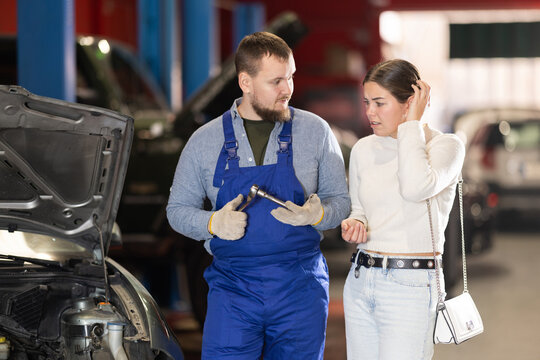 Male mechanic advises young woman client on repairing under hood of car in car service station
