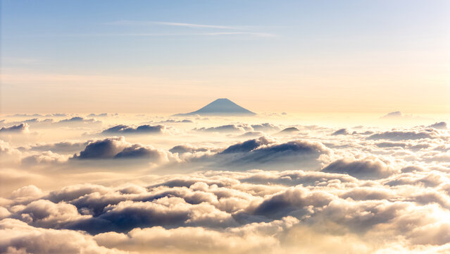 Aerial view of a mountain peak rising above a dense layer of clouds at sunrise, with soft golden light illuminating the sky and cloud formations in Dieng Kulon, Jawa Tengah, Indonesia.