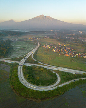 Aerial view of a highway interchange loop ramp surrounded by lush green rice paddies and rural settlements with a large mountain in the background in Indonesia.