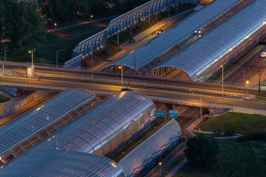Aerial view of the Trasa Torunska highway interchange featuring modern glass noise barriers and illuminated roads at dusk in Warsaw, Mazowieckie, Poland.