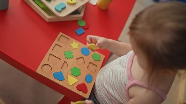 Toddler girl placing colorful wooden shape puzzle pieces into a peg board on a red table in a playroom; concentration.