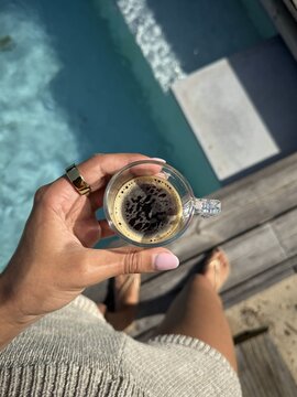 Hand holding an espresso above a swimming pool, capturing a peaceful morning ritual in a luxury holiday setting with natural light and summer vibes.