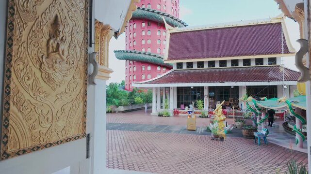 A person in white walks through the courtyard of Wat Sam Phran Buddhist temple in Bangkok, Thailand. The temple features ornate carvings and a unique pink tower