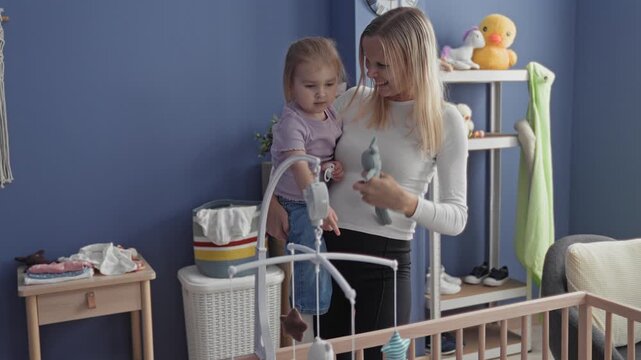 Mother holding toddler by crib, girl with finger to mouth and stuffed toy visible in nursery building; quiet tenderness.