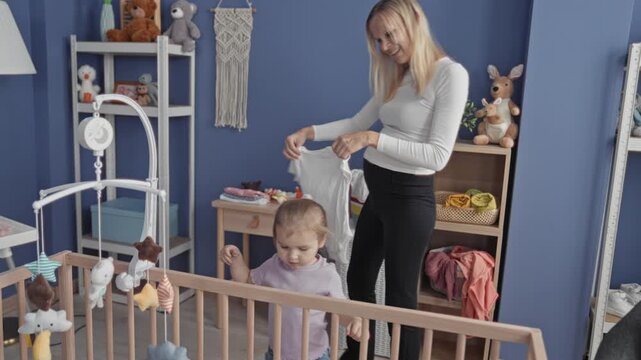 Woman handing a shirt to her little daughter at a wooden crib in a home building, reaching over a mobile and smiling; tenderness bonding.