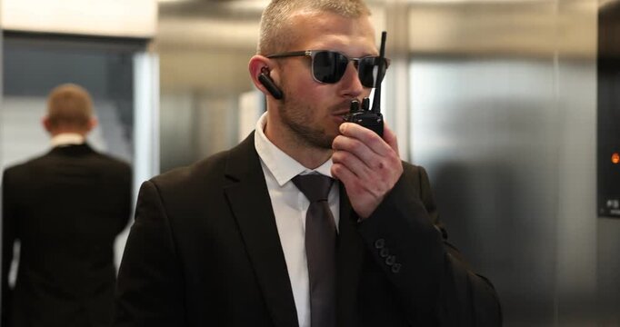 Bodyguard in suit with earpiece using walkie talkie while walking out from elevator indoors