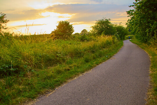 Aerial view of a paved path winding through a grassy field at sunset with trees lining the side and a person walking in the distance, Dewsbury, Dewsbury, United Kingdom.