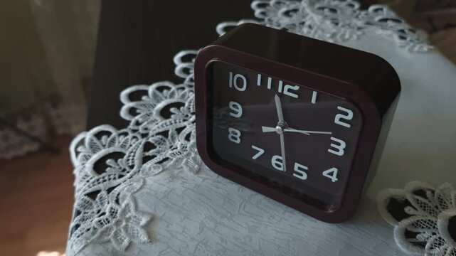 A square brown alarm clock rests on a white lace doily on a bedside table in the morning. The smooth camera pan highlights the domestic comfort of a slow morning routine.