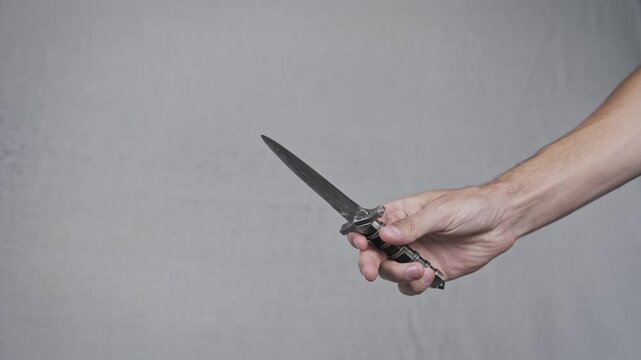 Close-up of a male hand firmly gripping and brandishing a vintage dagger against a neutral background. The sharp metal blade suggests concepts of aggression and historical combat.