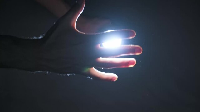 A male hand reaches towards a blindingly bright beam of light in a dark room with floating dust. Concept of spiritual awakening, divine connection, mystery, and finding hope.