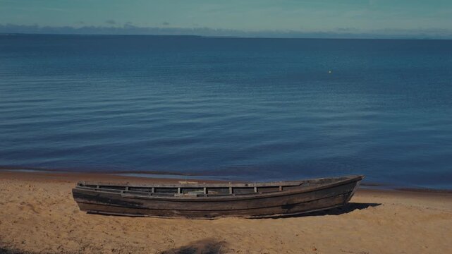 Wooden rowboat rests on golden sand. Calm blue water stretches to distant horizon. Clear sky frames peaceful coastal scene. Ideal for travel or nature content. No people present
