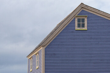 Obraz premium A side view of a vintage heritage purple wooden gabled roof cottage with cream colored trim around the eave and windows. The four-pane window sash frames are for a farmhouse. The sky is cloudy blue.