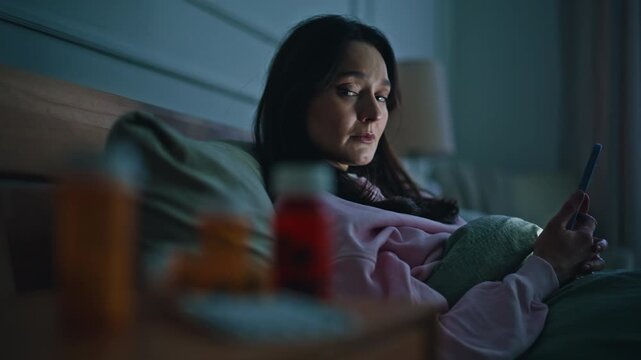 Sick woman watching cellphone lying evening bedroom closeup. Numerous drugs