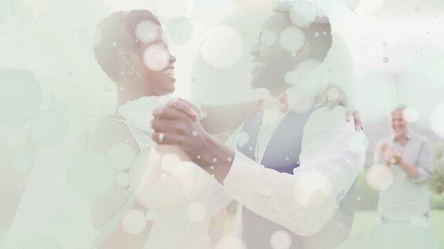Bokeh overlays thinning, newlyweds dancing on lawn, groom guiding bride, guests applauding wedding
