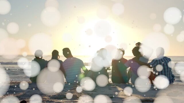 7 adults sitting beach facing sunset, bokeh thinning clearing group as one raising phone for travel