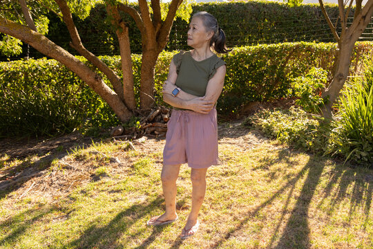 Senior Asian woman standing on sunny lawn crossing arms wearing smartwatch near branch and hedge