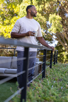 African American man holding ceramic mug and leaning on metal cable railing on deck