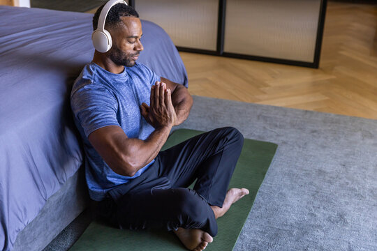 African American man meditating on green yoga mat beside bed, wearing blue shirt and white headset