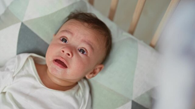 Hispanic baby boy holds wooden teether to mouth while lying on a patterned crib sheet inside a building crib; curiosity teething comfort.