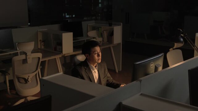 A businessman sitting alone at a dark office desk late at night looking at a computer monitor with a thoughtful expression.