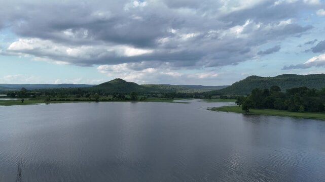 Drone view of Huai Sa Thot reservoir with lush green mountains under a cloudy blue sky in Kalasin, Thailand.