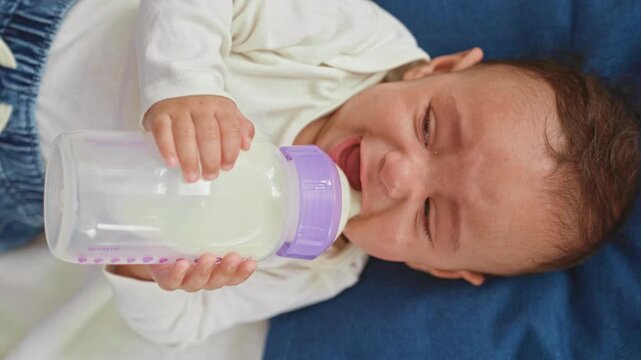 Hispanic baby boy holds milk bottle and cries with closed eyes and tiny hands while feeding on a blanket in a building; upset comfort.