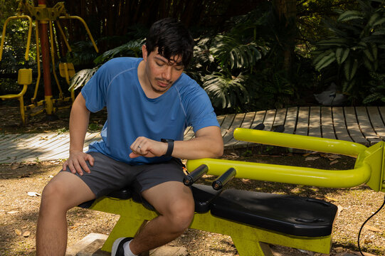 Man sitting on outdoor fitness equipment checking his smartwatch in a park surrounded by nature. Concept of physical activity, technology, and well being