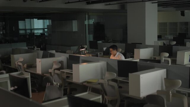 A lone office worker sitting at a workstation in a dark empty office space late at night with soft light from a computer screen.