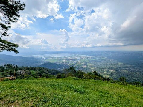 Scenic Mountain Landscape With Lush Green Grass Field Overlooking A Vast Valley And Lake Under A Cloudy Sky