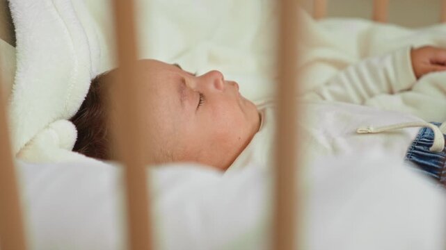 Baby boy sleeping in crib with cheek visible on white blanket inside a building; calm peaceful rest.