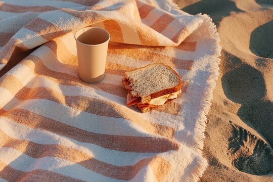 Striped beach towel with a paper cup and sandwich resting on sandy surface, evoking a relaxed outdoor dining experience by the shore