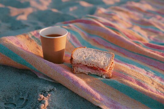 Striped beach towel with a paper cup of beverage and a sandwich placed on sandy surface, evoking a relaxed outdoor dining experience by the shore